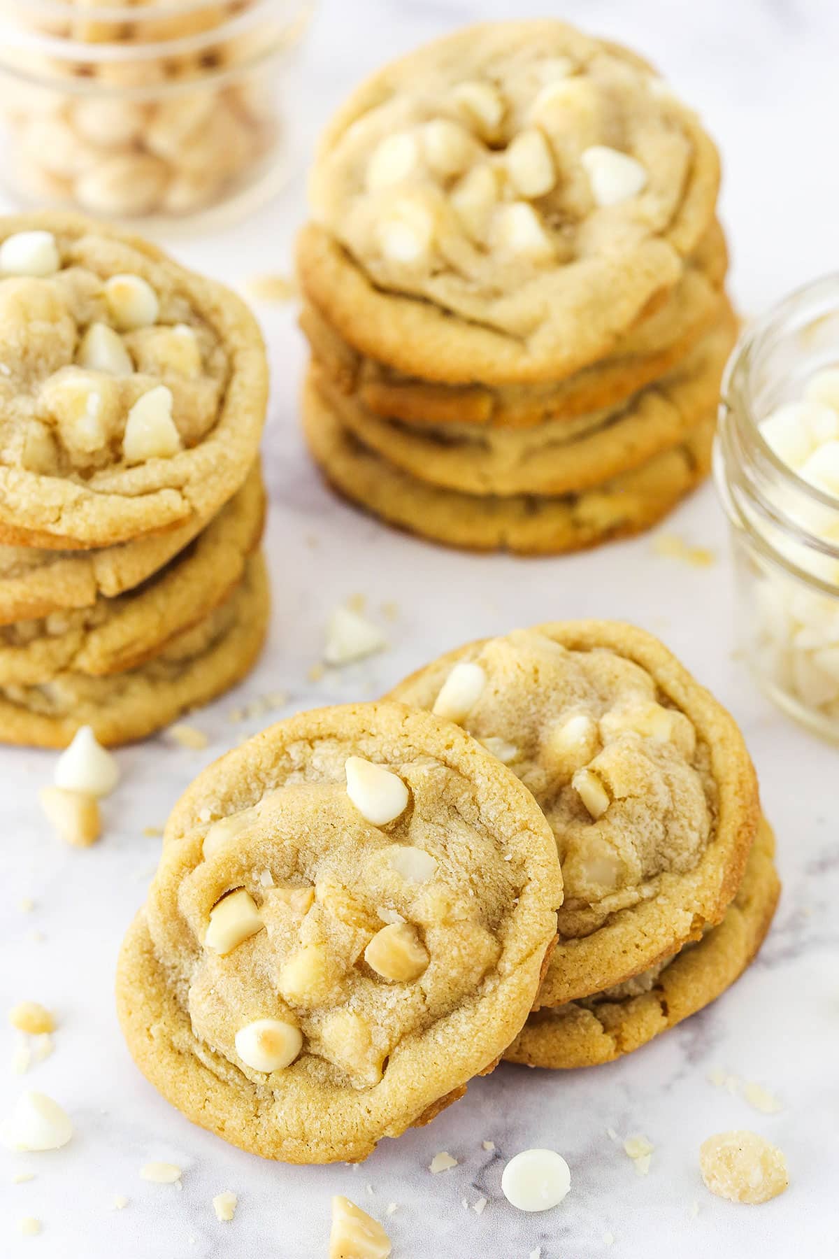 White Chocolate Macadamia Nut Cookies stacked on a marble table top with white chocolate chips and macadamia nuts in the background