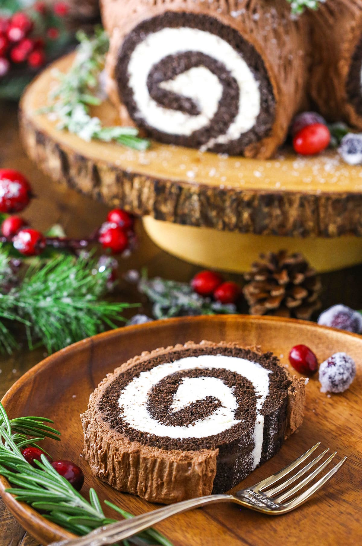 A slice of Yule Log Cake on a wooden plate with sugared cranberries and rosemary sprigs and a fork with a Yule Loge Cake in the background