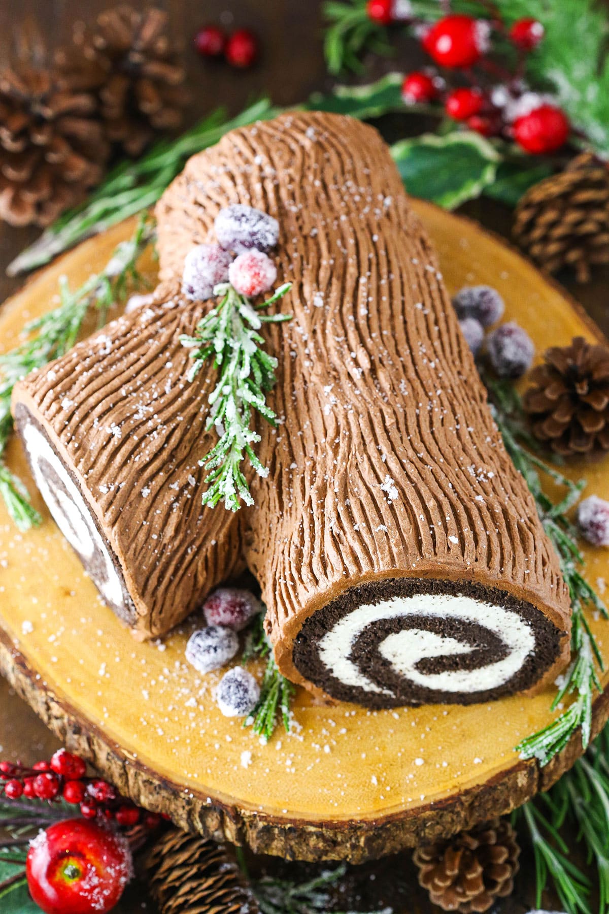 Overhead view of a Yule Log Cake decorated with sugared cranberries and rosemary sprigs on a wooden cake stand