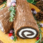 Side view of a Yule Log Cake decorated with sugared cranberries and rosemary sprigs on a wooden cake stand