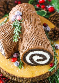 Side view of a Yule Log Cake decorated with sugared cranberries and rosemary sprigs on a wooden cake stand