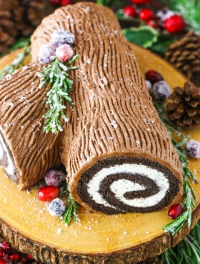 Side view of a Yule Log Cake decorated with sugared cranberries and rosemary sprigs on a wooden cake stand