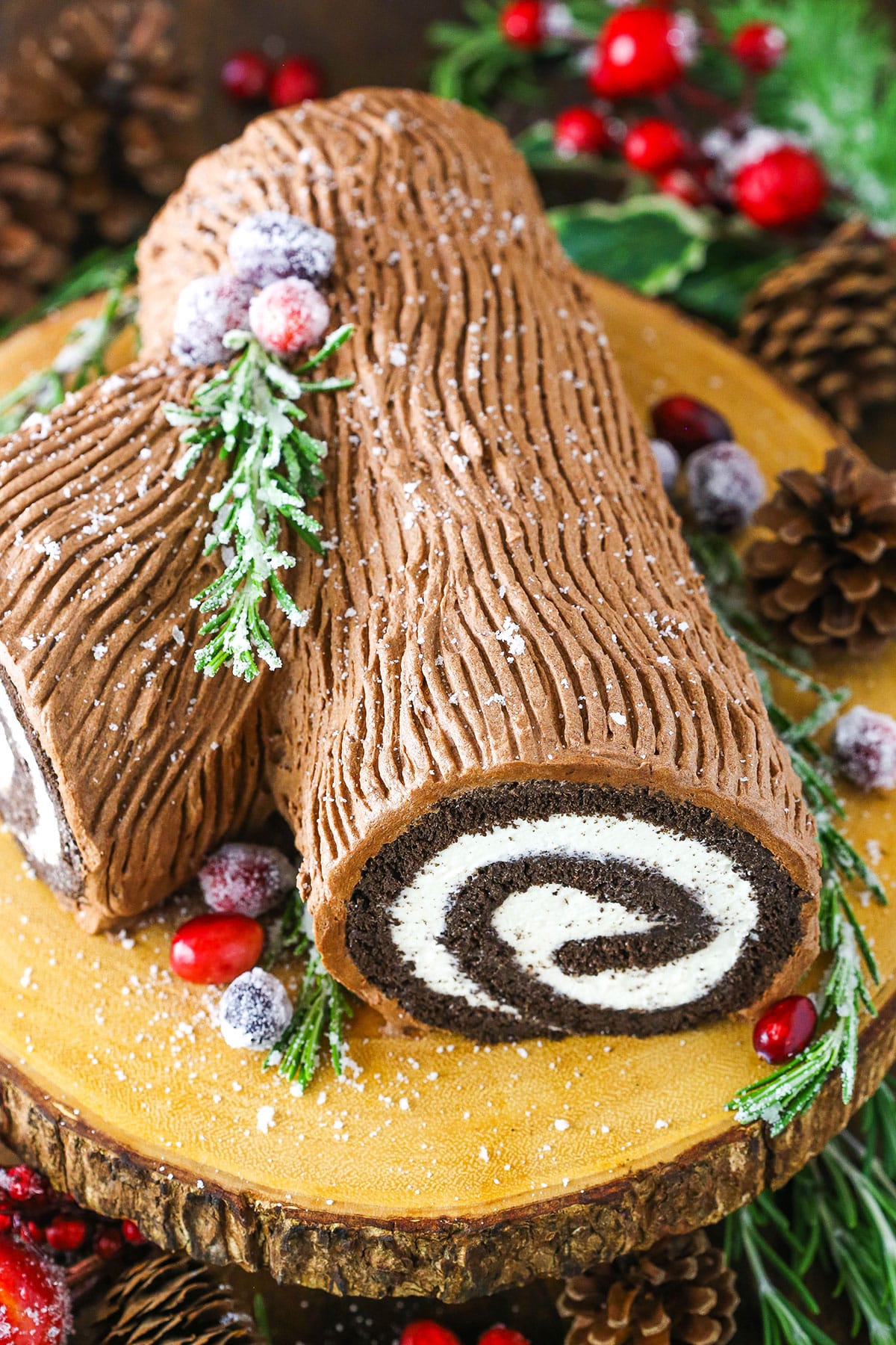 Side view of a Yule Log Cake decorated with sugared cranberries and rosemary sprigs on a wooden cake stand