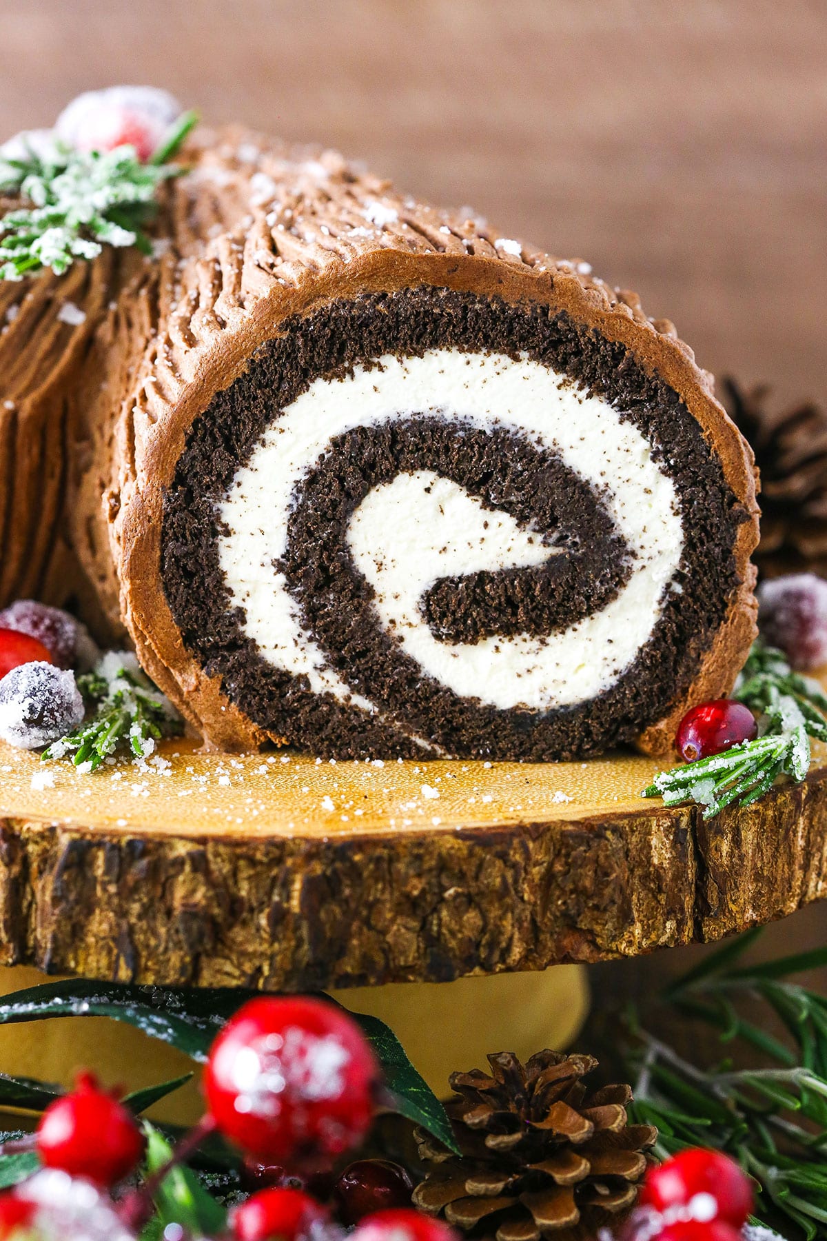 Side view of a Yule Log Cake decorated with sugared cranberries and rosemary sprigs on a wooden cake stand