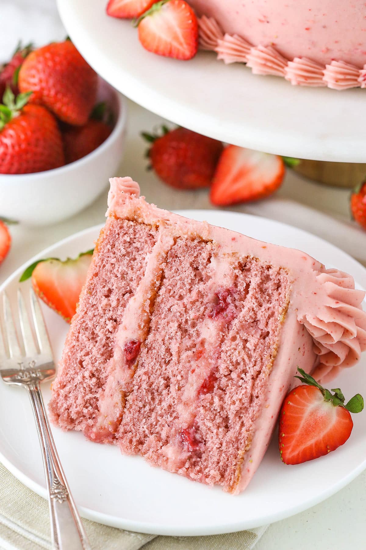 A slice of strawberry cake on a plate with a fork near fresh strawberries.