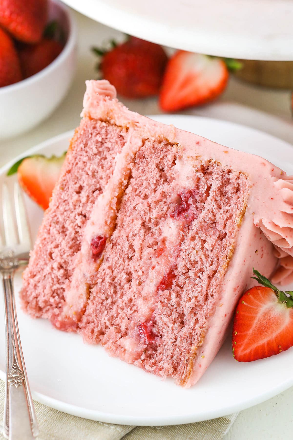 From batter to frosting, this 3-layer strawberry cake is absolutely brimming with mouthwatering strawberry flavor from real fruit. Yum! A slice of strawberry cake on a plate with a fork near fresh strawberries.