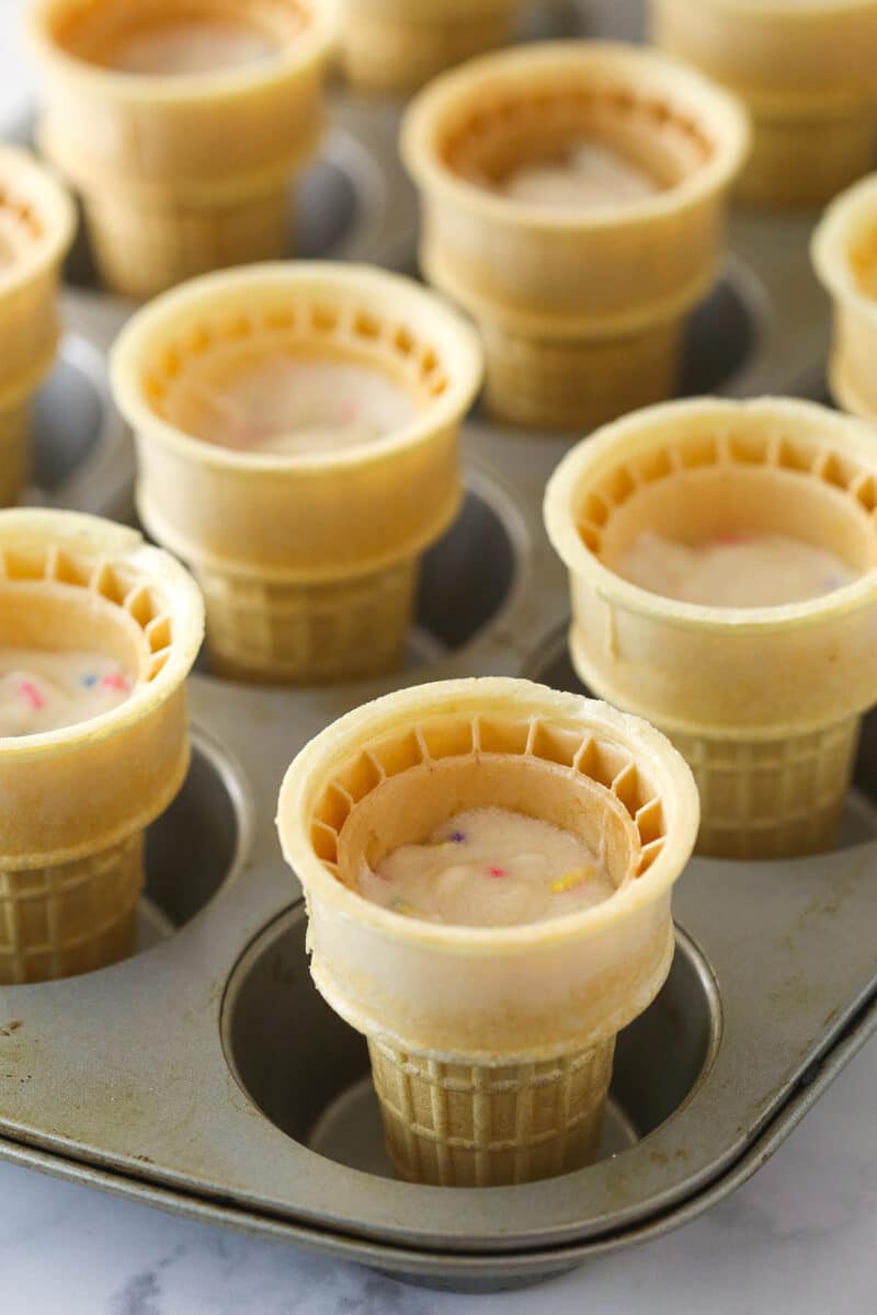 Ice cream cones filled with cupcake batter standing in cupcake tins ready to be baked.