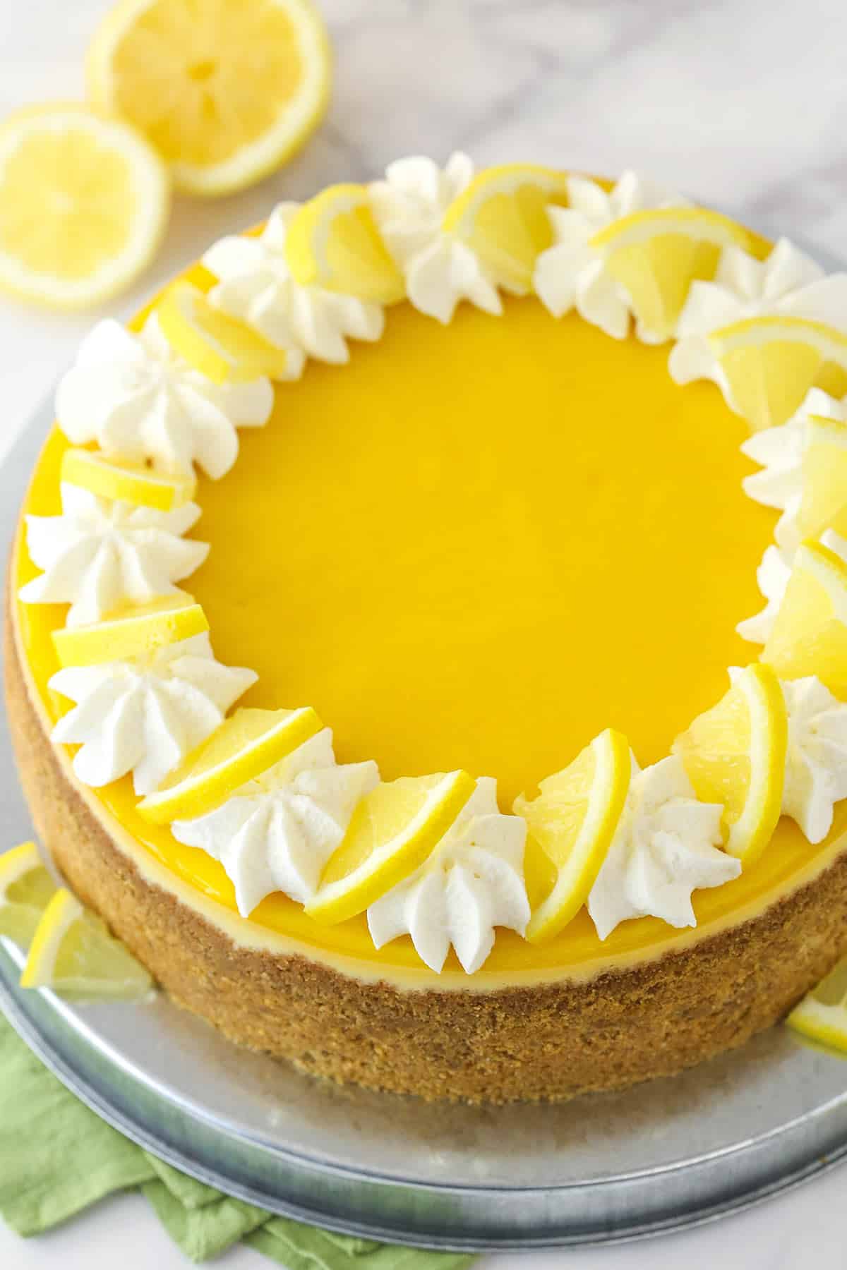 Lemon cheesecake is perfect for summer. Creamy lemon cheesecake filling is baked into sweet Graham cracker crust and topped with lemon curd. A whole lemon cheesecake on a serving plate.