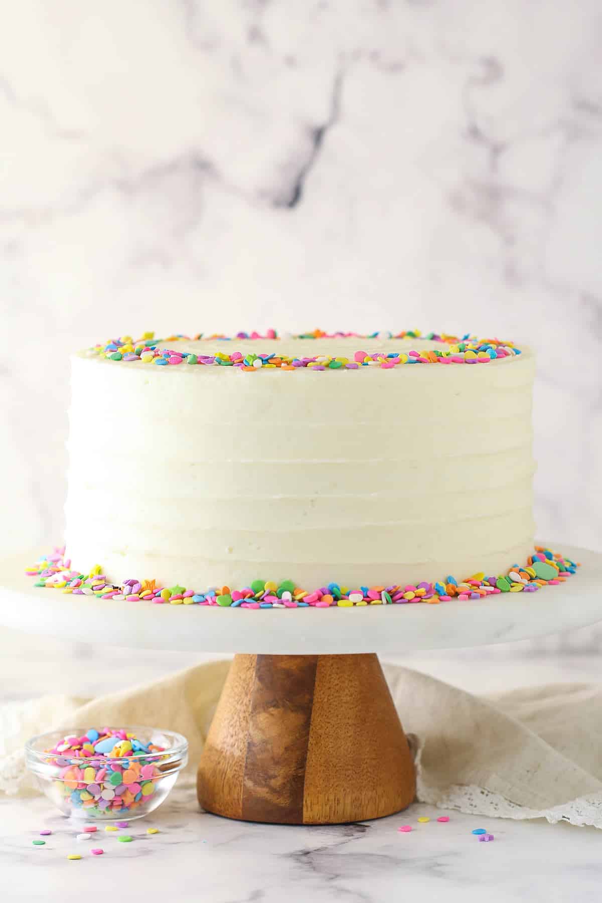 Moist vanilla layer cake on a cake stand near a bowl of sprinkles.