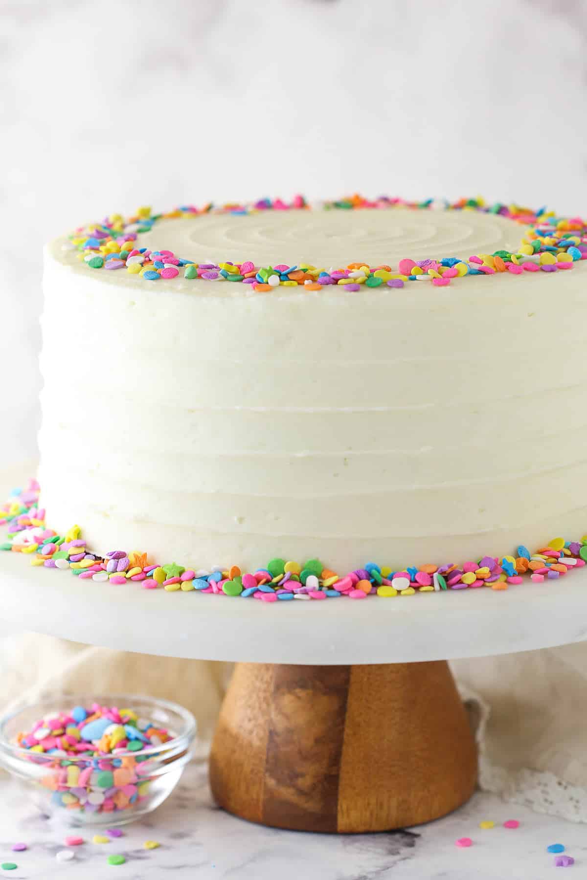 Moist vanilla layer cake on a cake stand near a bowl of sprinkles.