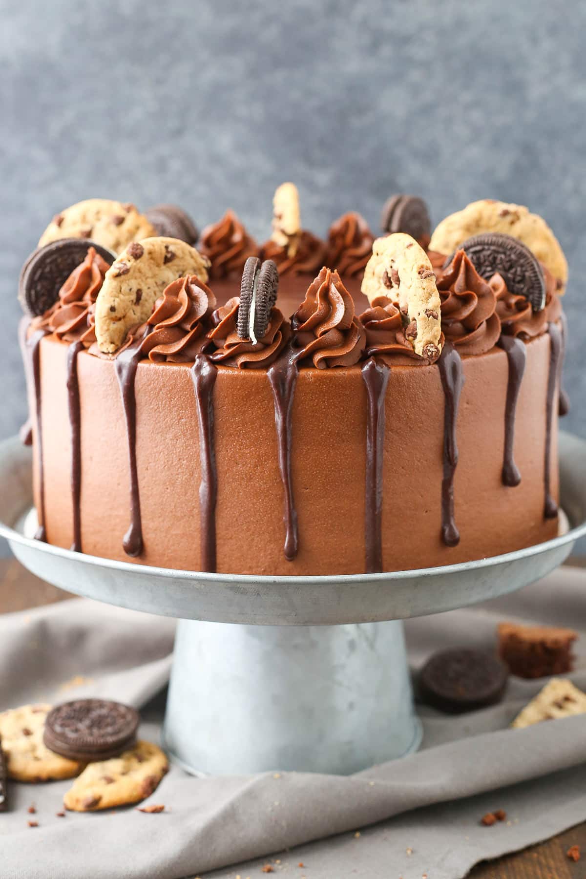 Oreo brookie cake on a cake stand surrounded by Oreos and chocolate chip cookies.