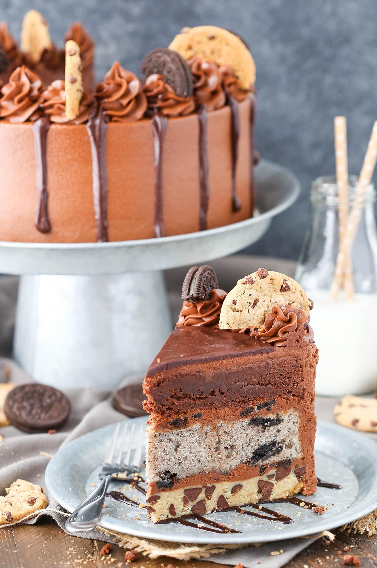 A slive of brookie cake on a plate with a fork in front of a whole Oreo brookie layer cake near a carafe of milk.