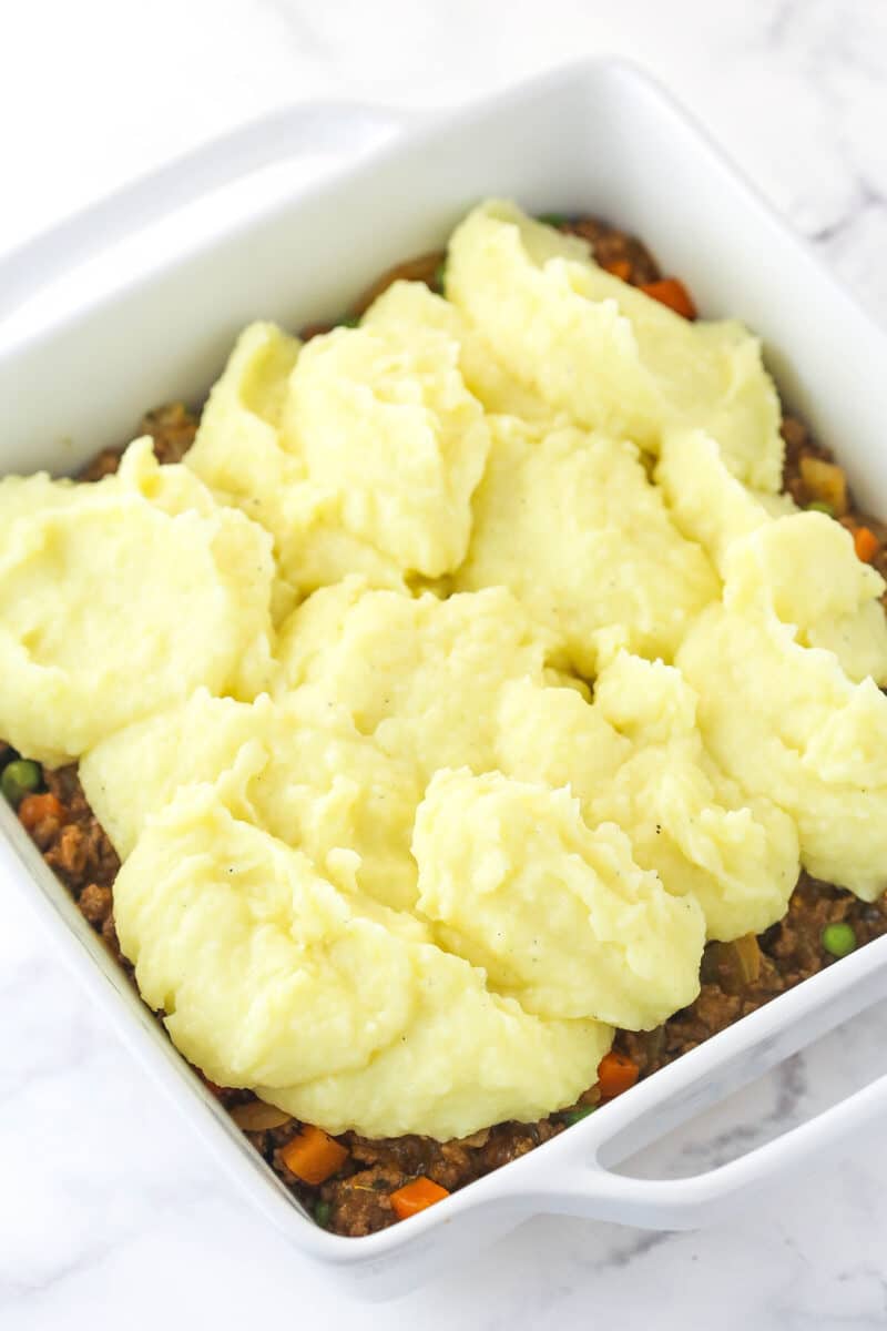 This easy Sheperd's Pie is made of perfectly seasoned ground beef and veggies topped with mashed potatoes and cheese and baked to bubbly. Scooping mashed potatoes over a meat mixture in a baking dish.