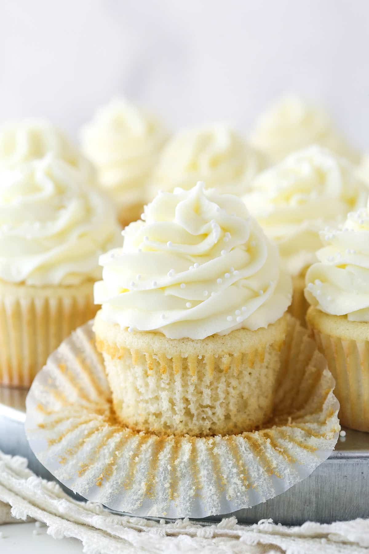 This white cupcake recipe produces moist, fluffy vanilla cupcakes topped with vanilla buttercream. They are sure to become a new favorite. White cupcakes on a serving tray. One is half way unwrapped.
