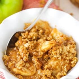 A single serving of Apple Crisp in a white bowl with a spoon with apples in the background