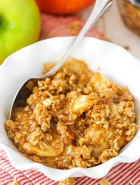 A single serving of Apple Crisp in a white bowl with a spoon with apples in the background