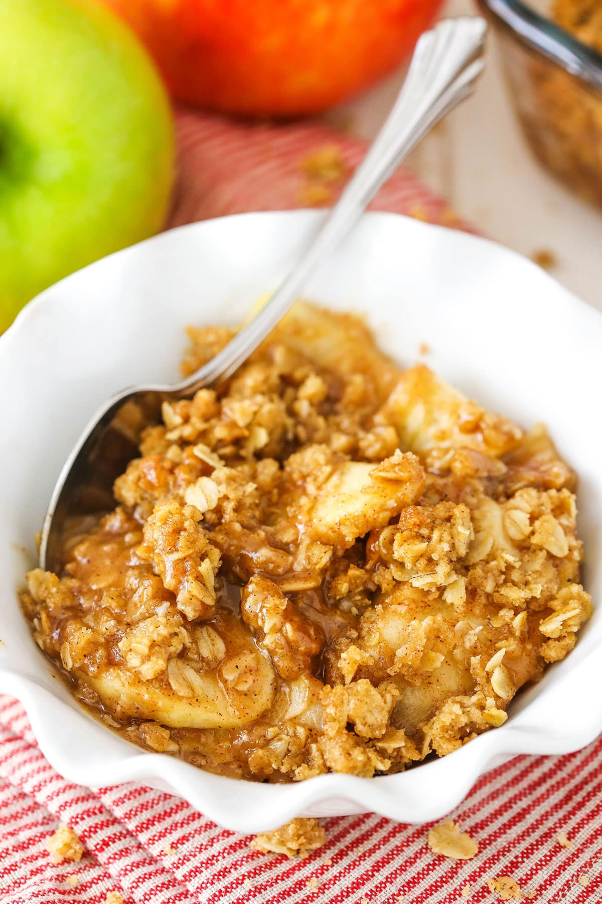 A single serving of Apple Crisp in a white bowl with a spoon with apples in the background