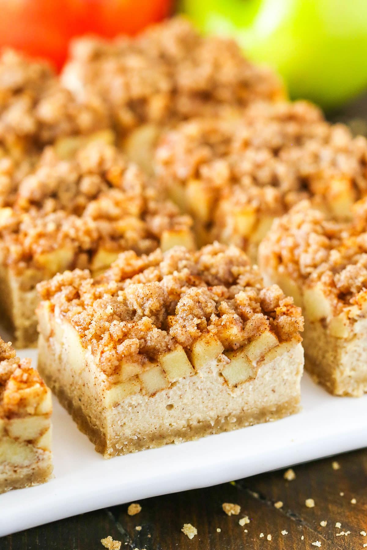 These Apple Streusel Cheesecake Bars are a delightful apple recipe perfect for Thanksgiving dessert. They have a buttery shortbread crust, creamy cheesecake filling and are topped with cinnamon apples and a cinnamon streusel! Apple Streusel Cheesecake Bars on a white platter with apples in the background