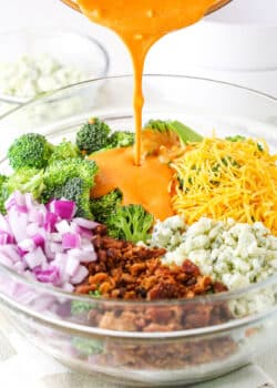 Pouring buffalo sauce over Buffalo Broccoli Salad ingredients in a clear glass bowl on a white table