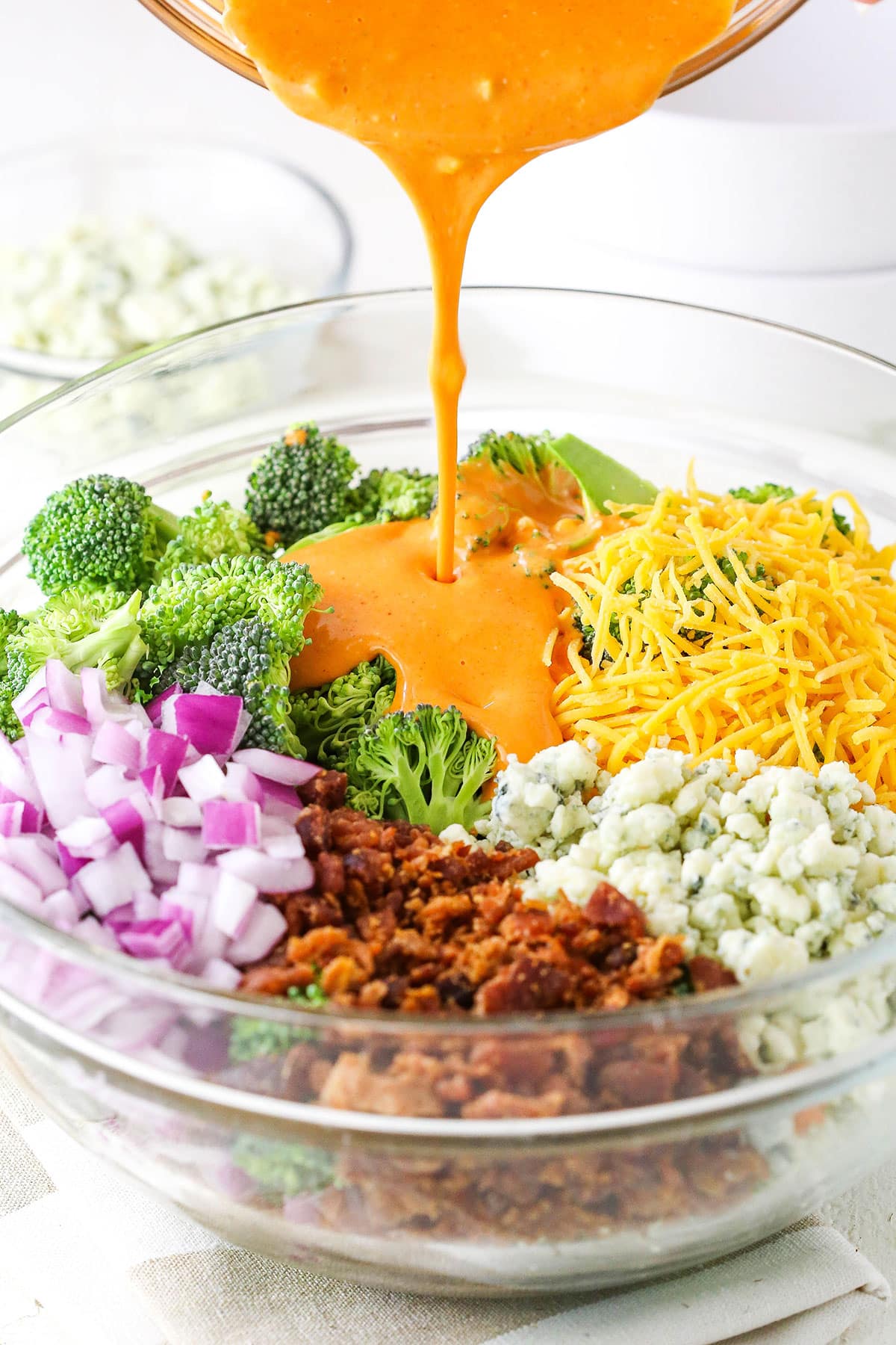 Pouring buffalo sauce over Buffalo Broccoli Salad ingredients in a clear glass bowl on a white table