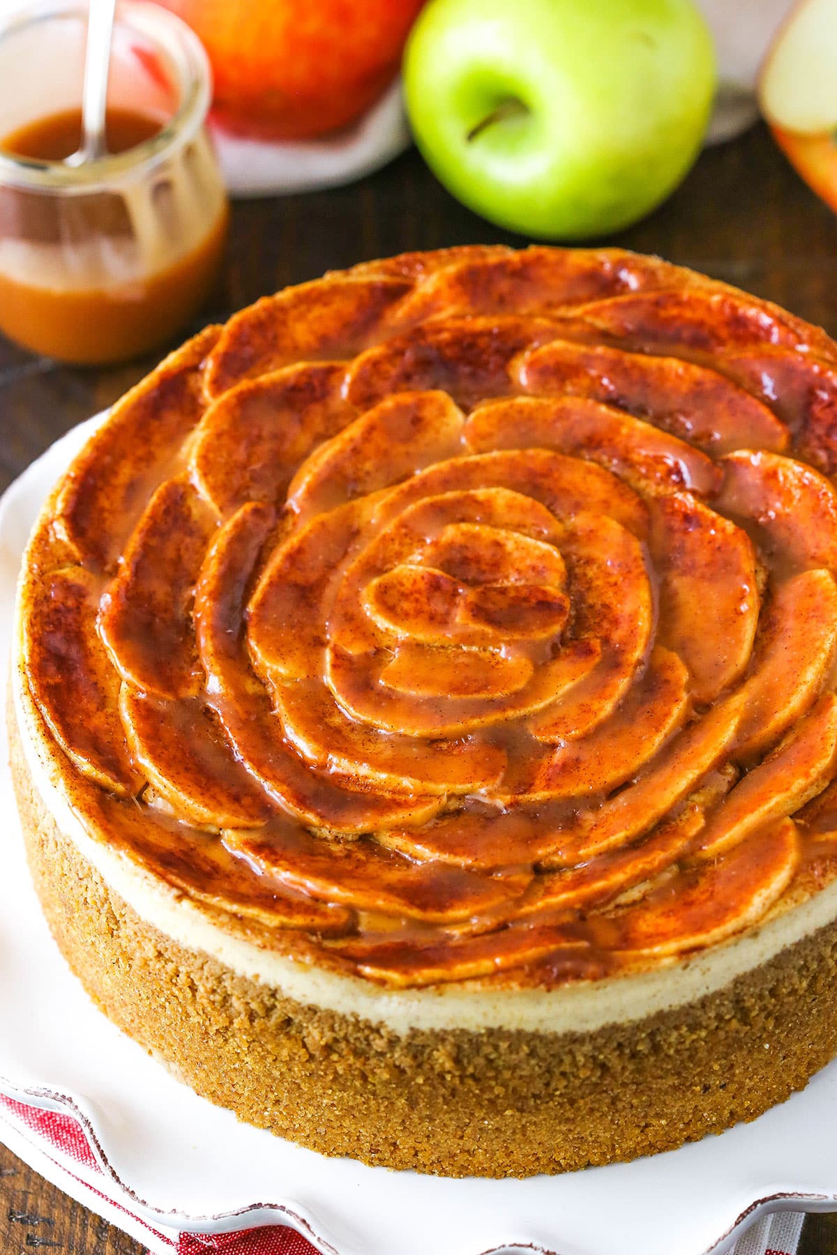 A full Caramel Apple Cheesecake on a white cake stand with caramel and apples in the background