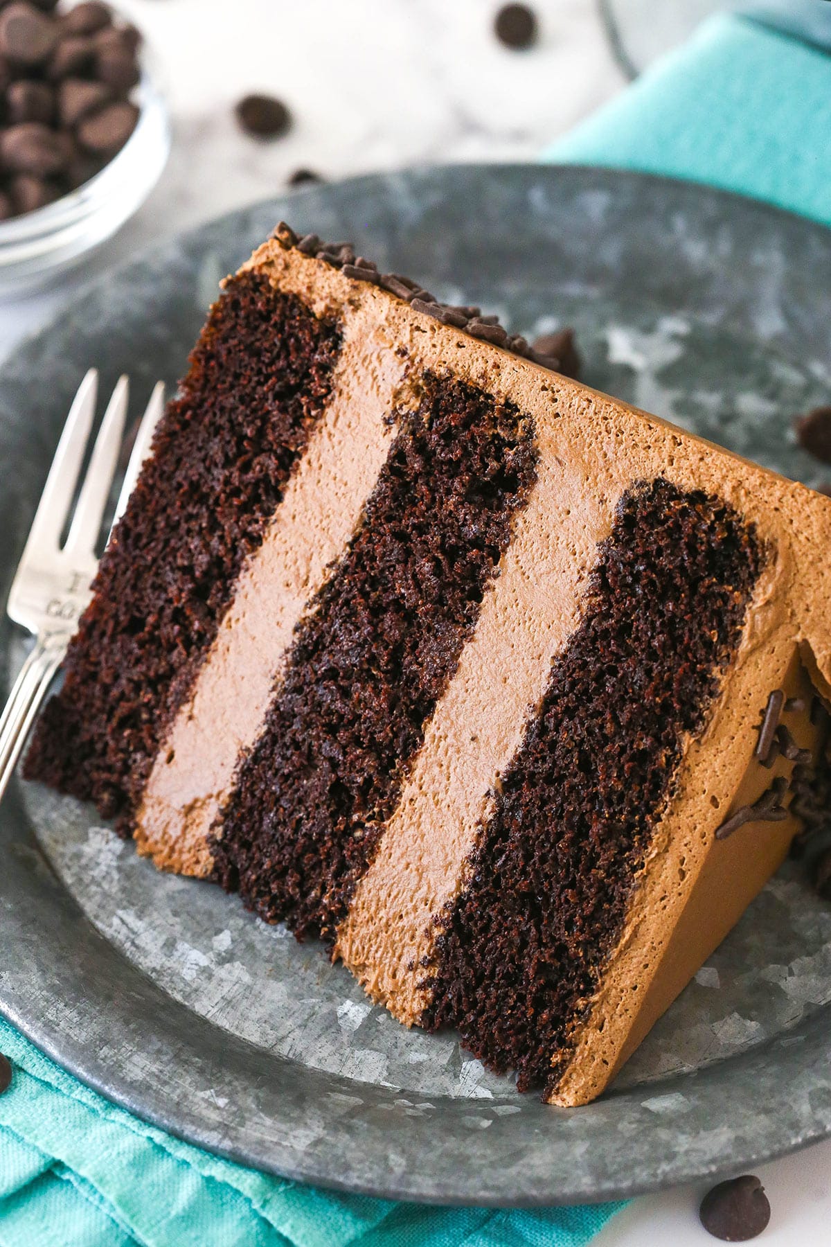 A slice of chocolate mousse cake on a plate with a fork near a bowl of chocolate chips.