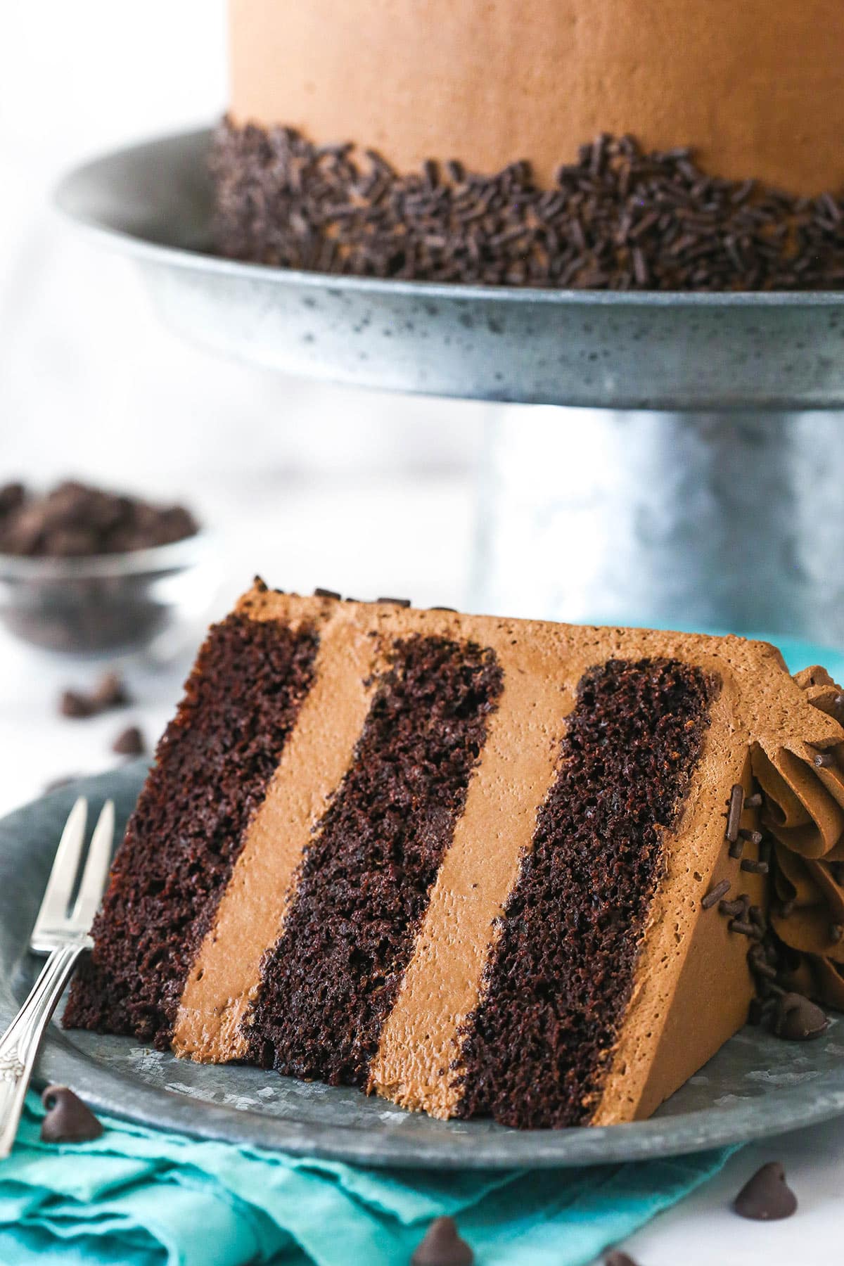 A slice of chocolate mousse cake on a plate with a fork near a whole chocolate mousse cake on a cake stand and a bowl of chocolate chips.