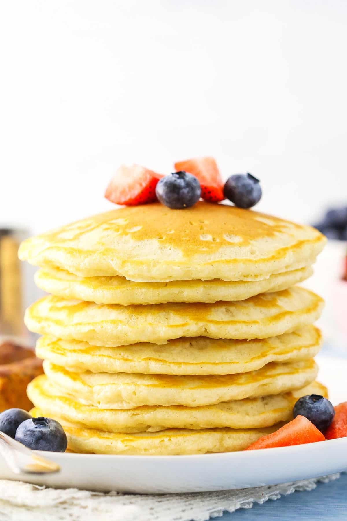 Side view of seven Fluffy Homemade Pancakes stacked on a white plate and topped with cut strawberries and blueberries
