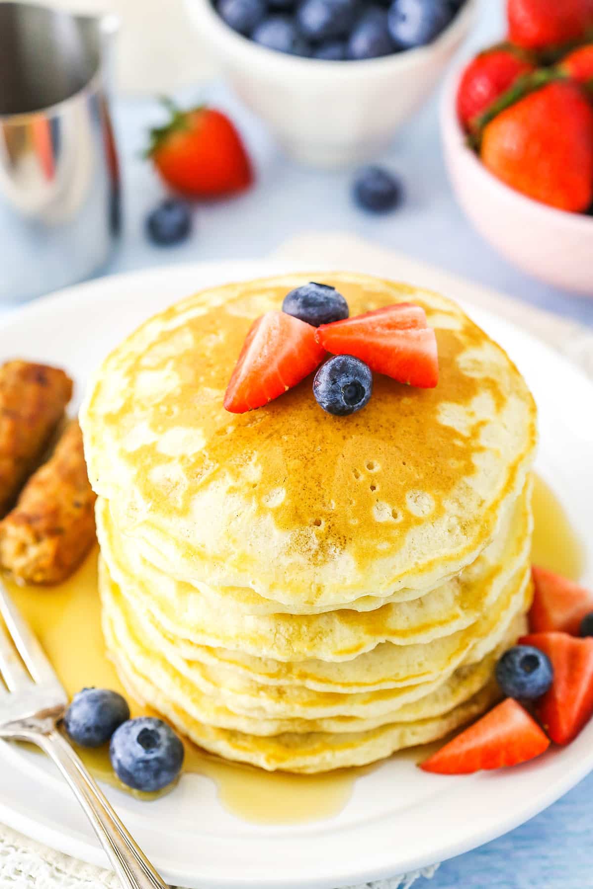 Pancakes stacked next to a fork, sausage links and cut strawberries on a white plate