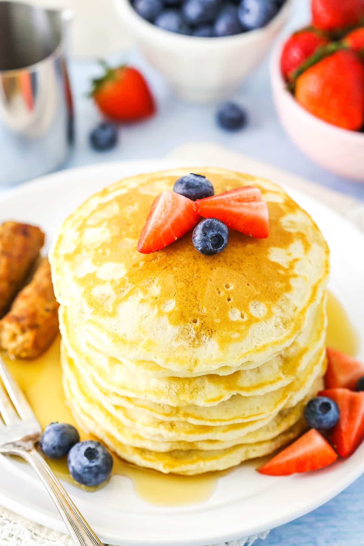 Fluffy Homemade Pancakes stacked next to a fork, sausage links and cut strawberries on a white plate