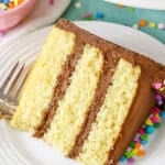 A slice of Yellow Cake with Chocolate Frosting next to a fork on a white plate