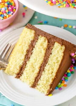 A slice of Yellow Cake with Chocolate Frosting next to a fork on a white plate