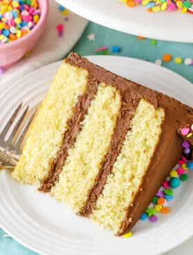 A slice of Yellow Cake with Chocolate Frosting next to a fork on a white plate