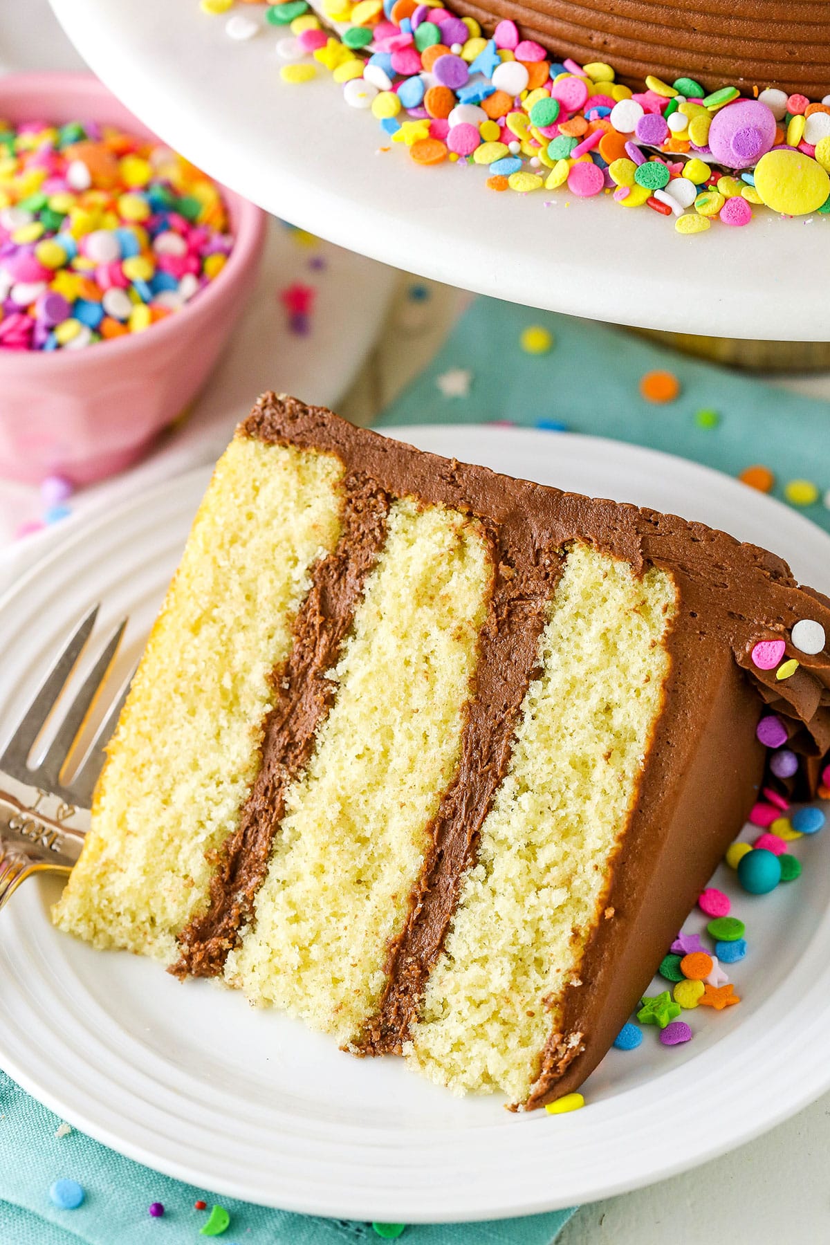 A slice of Yellow Cake with Chocolate Frosting next to a fork on a white plate
