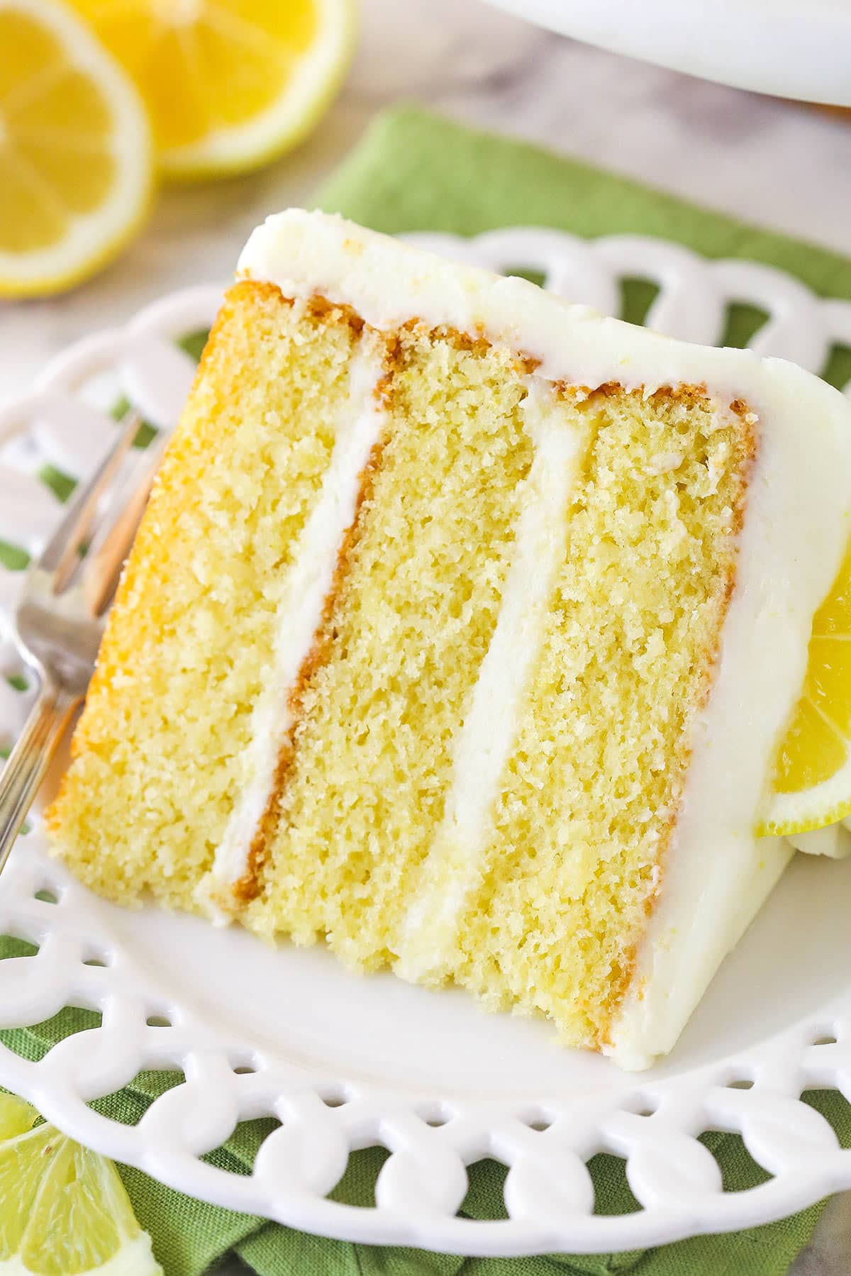 This lemon layer cake is made up of three layers of fluffy lemon cake that are filled and frosted with vibrant lemon cream cheese frosting. A slice of lemon layer cake on a plate.