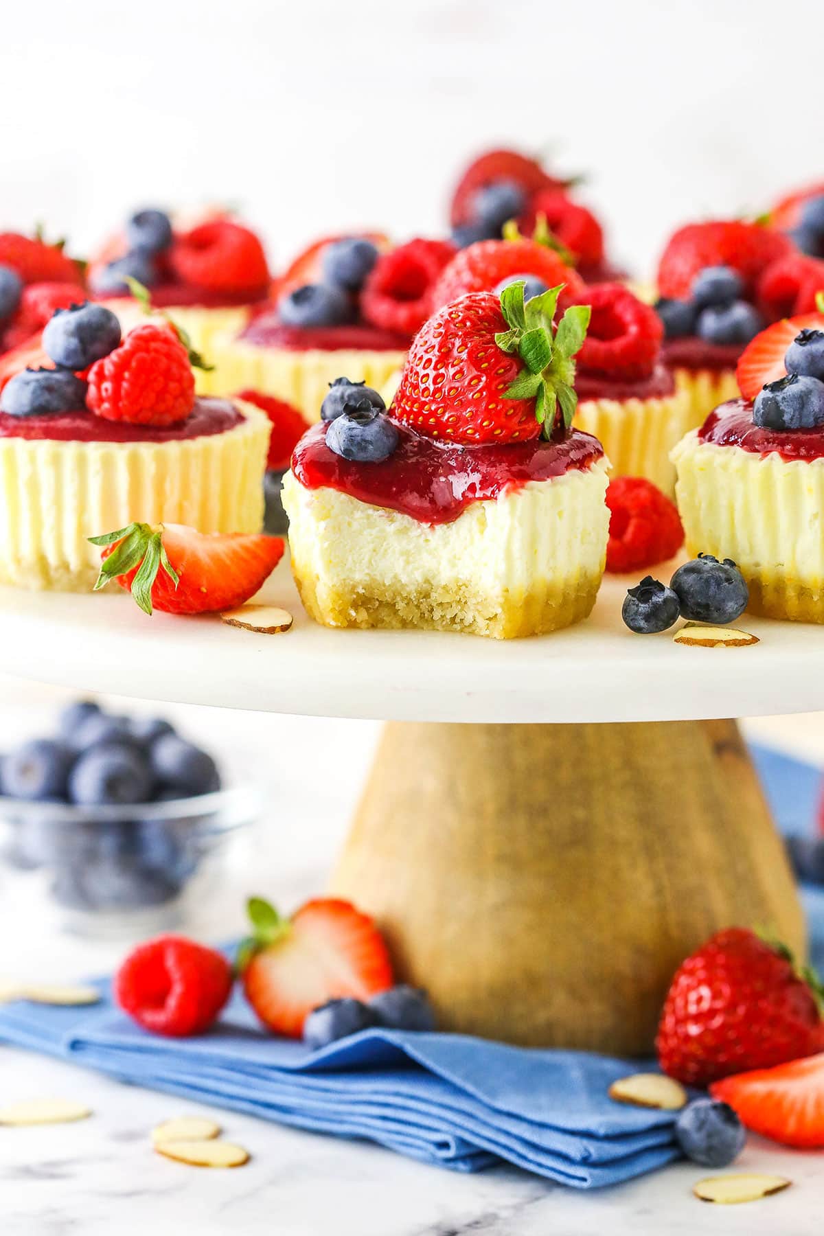 Whole Mini Berry Almond Cheesecakes with one missing a bite topped with strawberries, blueberries and raspberries on a white cake stand