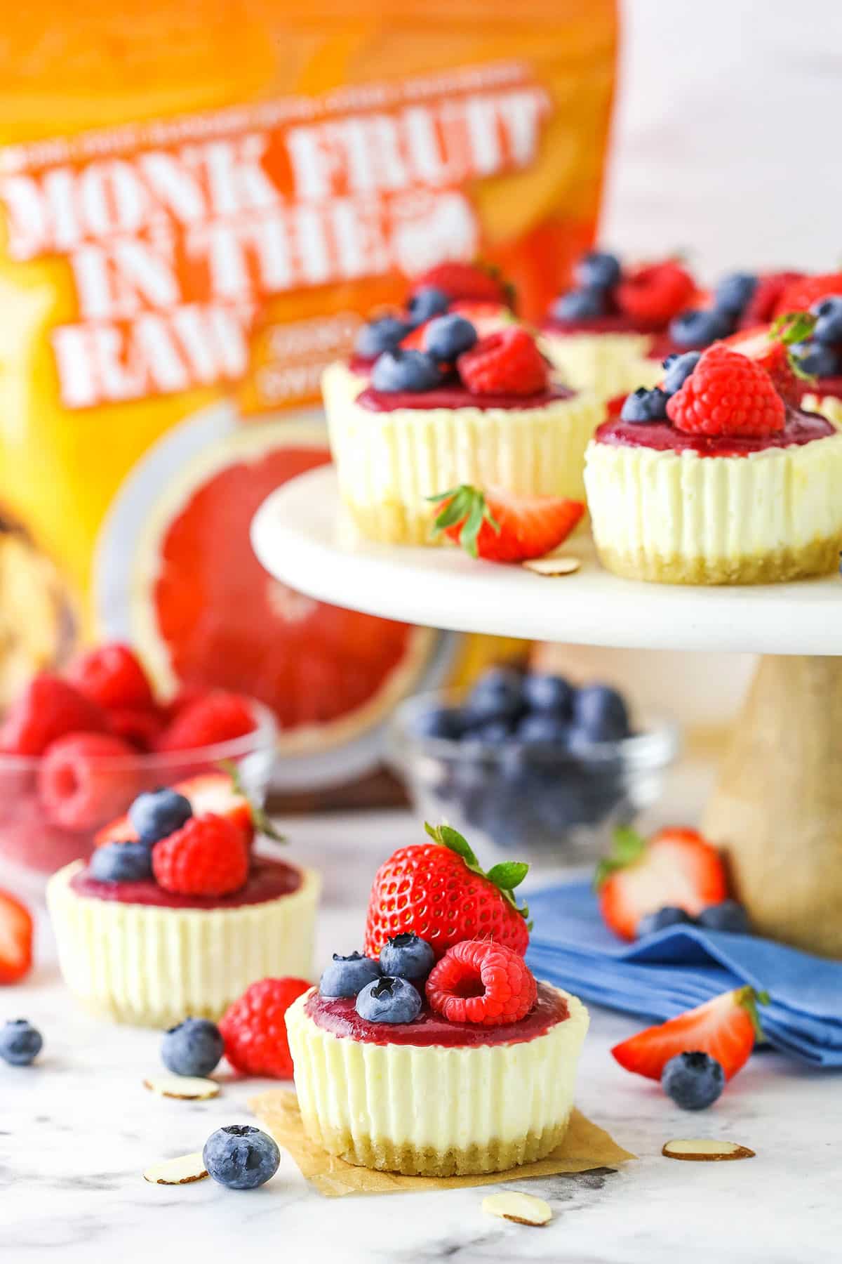 Mini Berry Almond Cheesecakes on a white cake stand with a bag of Monk Fruit In The Raw in the background