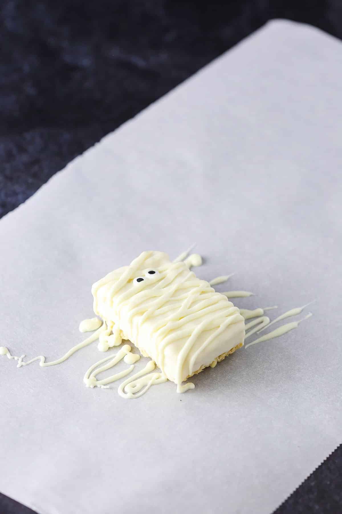 A step in making Mummy Rice Krispies Treats showing the completed treat decorated with eyes and piped white chocolate drying on parchment paper