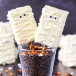 Two Mummy Rice Krispies Treats on orange and black straws in a glass filled with chocolate sprinkles