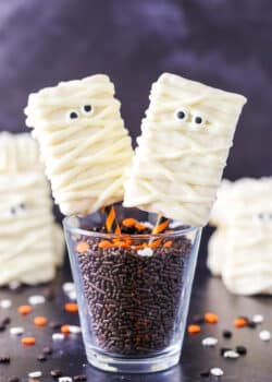 Two Mummy Rice Krispies Treats on orange and black straws in a glass filled with chocolate sprinkles