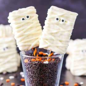 Two Mummy Rice Krispie Treats on orange and black straws in a glass filled with chocolate sprinkles.