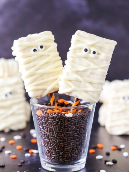 Two Mummy Rice Krispie Treats on orange and black straws in a glass filled with chocolate sprinkles.
