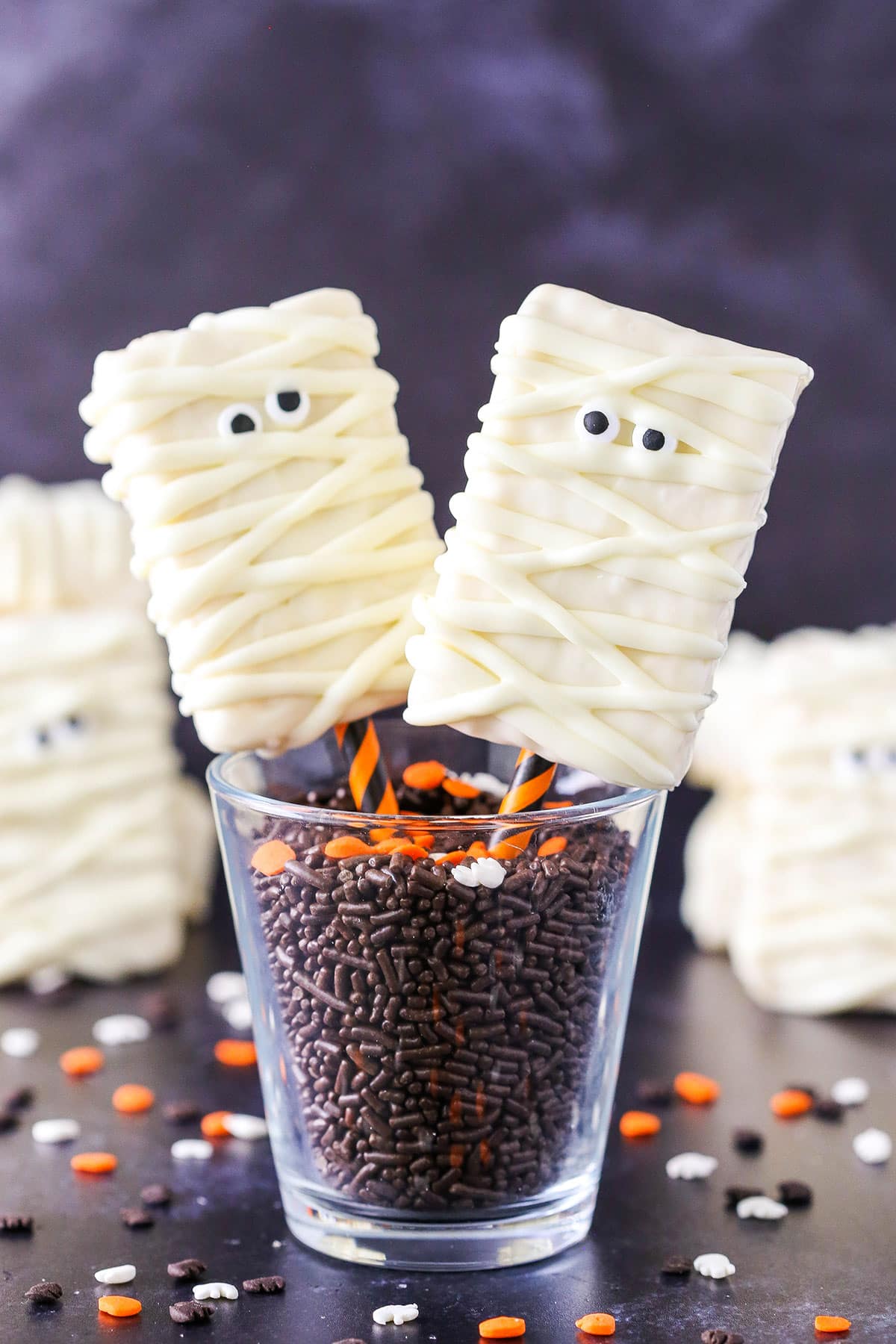 These Mummy Rice Krispie Treats are such a fun Halloween party food idea! Made with only three ingredients, they're the perfect easy recipe for Halloween parties. Two Mummy Rice Krispies Treats on orange and black straws in a glass filled with chocolate sprinkles