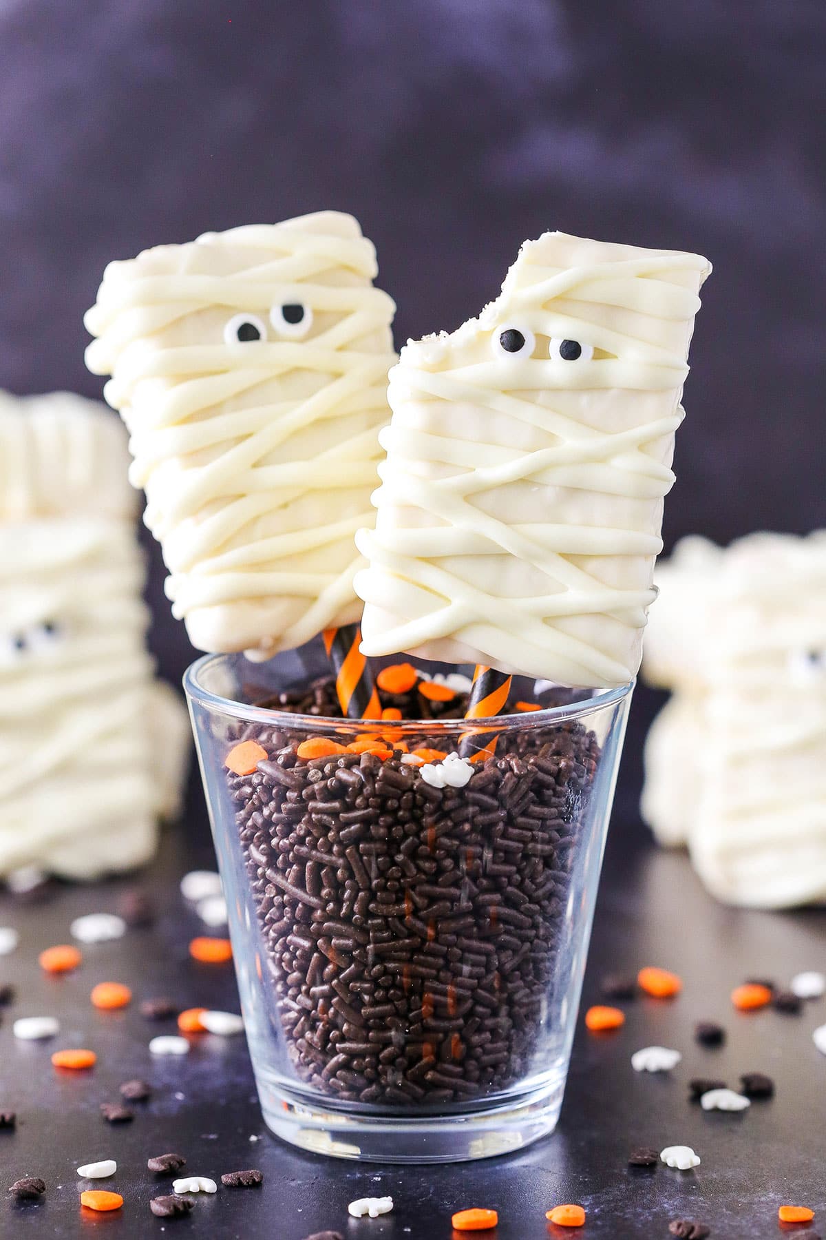 Two Mummy Rice Krispies Treats, one with a bite removed, on orange and black straws in a glass filled with chocolate sprinkles