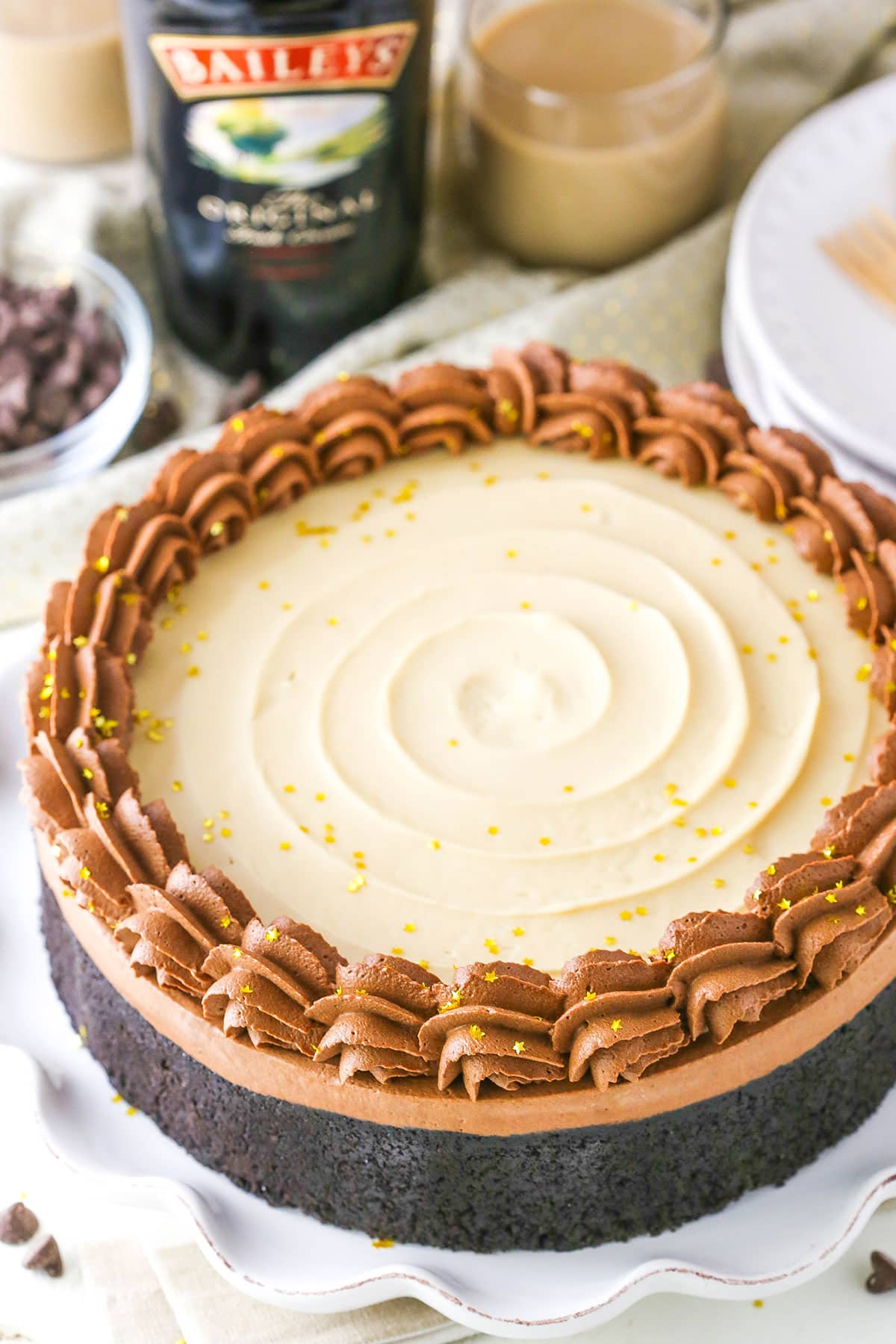 No Bake Baileys Chocolate Cheesecake on a white cake stand with a bottle and glass of Baileys in the background