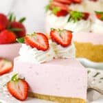 A slice of no bake strawberry cheesecake served on a plate with a fork near a full cheesecake and a bowl of fresh strawberries.