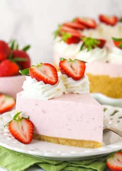 A slice of no bake strawberry cheesecake served on a plate with a fork near a full cheesecake and a bowl of fresh strawberries.