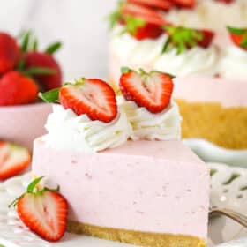 A slice of no bake strawberry cheesecake served on a plate with a fork near a full cheesecake and a bowl of fresh strawberries.
