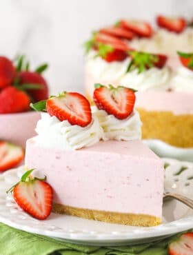 A slice of no bake strawberry cheesecake served on a plate with a fork near a full cheesecake and a bowl of fresh strawberries.