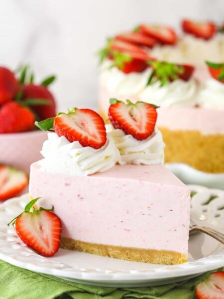 A slice of no bake strawberry cheesecake served on a plate with a fork near a full cheesecake and a bowl of fresh strawberries.